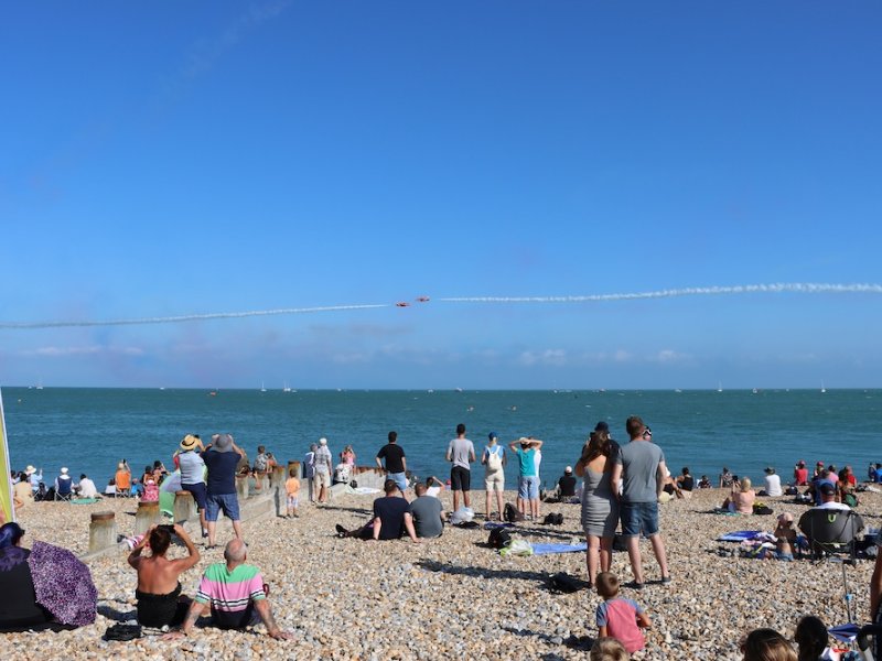 People on a shingle beach watching an air display over the sea
